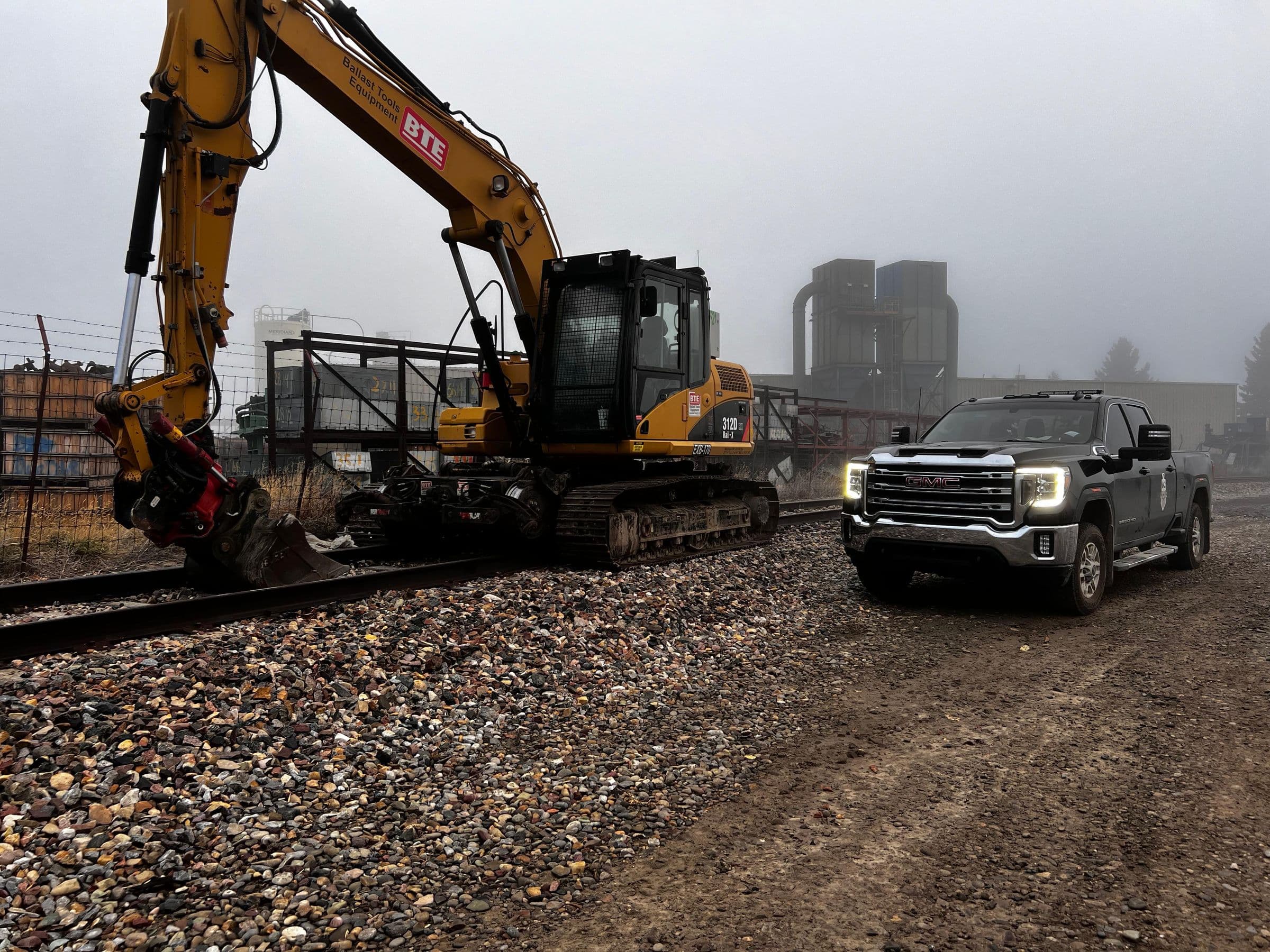 Excavator and pickup truck beside a ballasted track shoulder on a foggy day